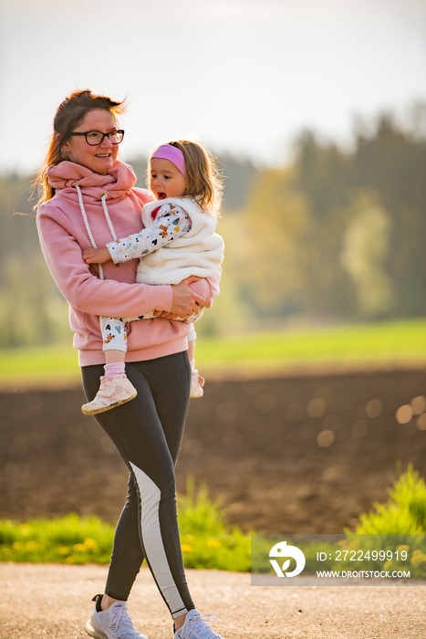 Mother and child walking on countryside road between agricultural fields towards vilage from forest.