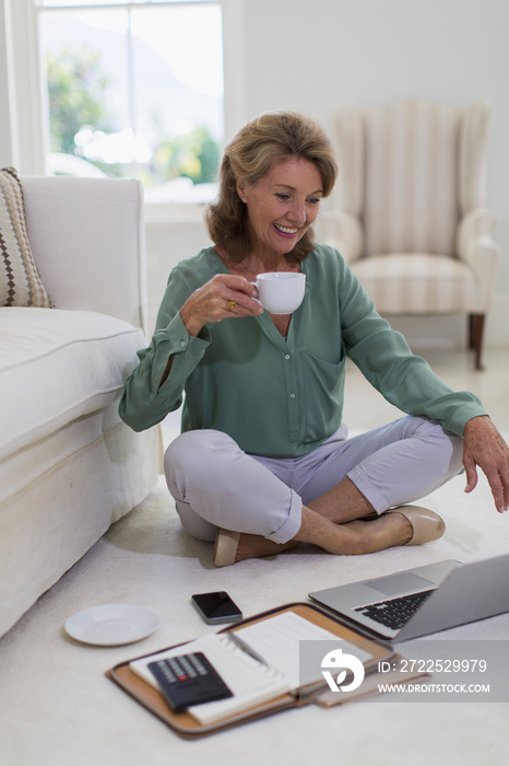 Senior woman drinking coffee at laptop on living room floor