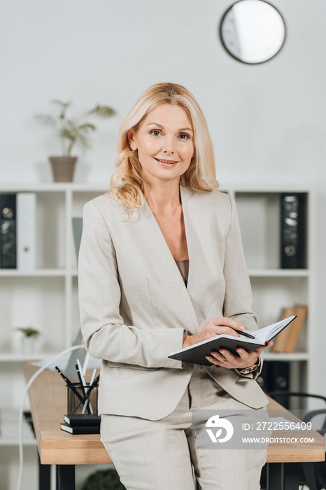 happy mature businesswoman writing in notepad and smiling at camera in office