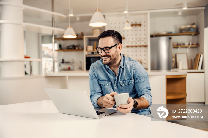 A happy man drinking coffee and looking at the laptop at his cozy home.