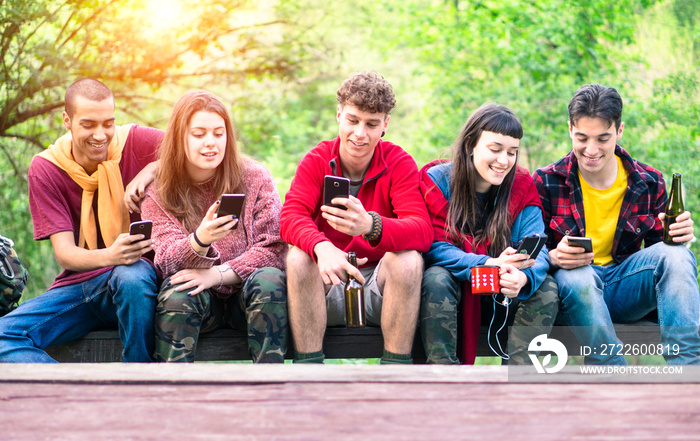 Group of young friends in a row holding mobile phone looking down smiling - Happy teenagers on hiking clothing  sitting outdoor using smartphone - Concept of teens friendship , technology addiction
