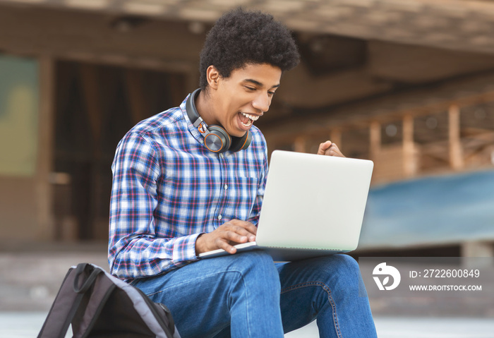 Excited young guy celebrating victory with laptop outdoors