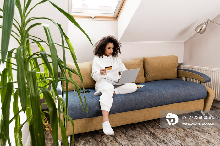 Selective focus of African American girl holding credit card near laptop while sitting on sofa