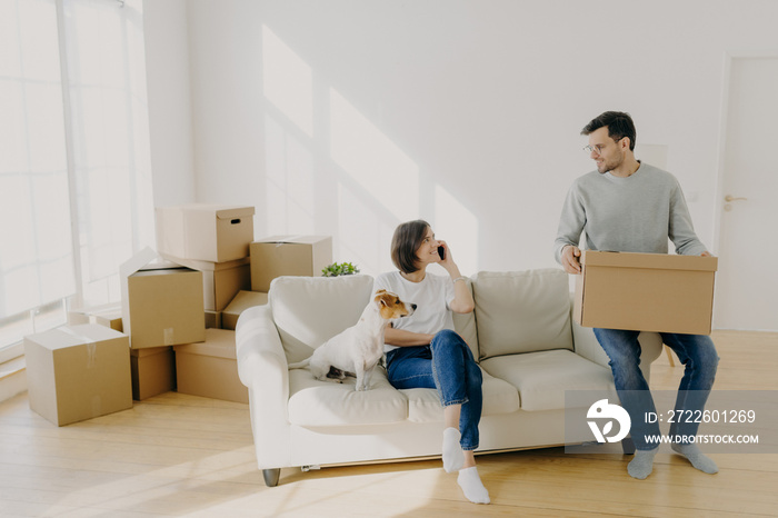 Photo of happy couple move in new home, pose on sofa with pet and boxes, have relocation, unpack boxes, female calls delivery service, carry belongings in carton containers. Welcome to new apartment