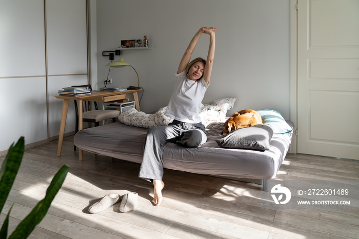 Middle-aged female in pajamas stretches out hands. Half-awake lady does warm-up stretching muscles on uncovered bed. Woman enjoys morning with dog in cozy bedroom starting day with lazy gymnastics