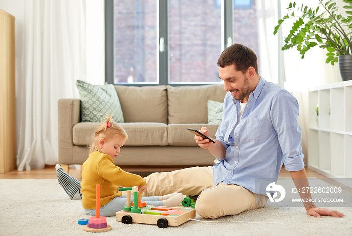 family, fatherhood and people concept - happy father with smartphone and little baby daughter playing with wooden toy toy blocks kit at home