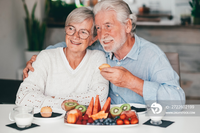 Lovely smiling senior couple at home having breakfast together with muffin, milk and fresh seasonal fruit, healthy eating concept