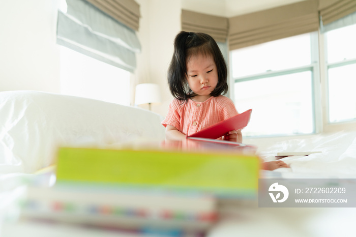 Selective focus of Asian toddler girl concentrate reading books foreground with pile of books on the bed in the natural morning sunlight, reading is knowledge, preschool education, child development.