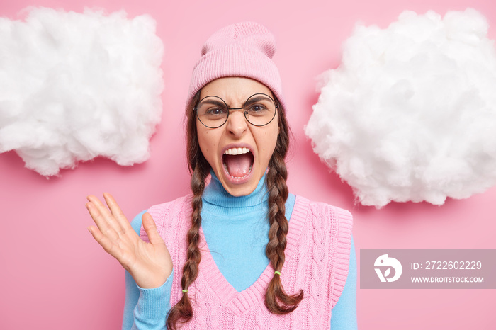 People negative emotions and feelings concept. Annoyed teenage girl exclaims from anger yells loudly keeps mouth opened has two pigtails wears hat round spectacles turtleneck with vest stands indoor