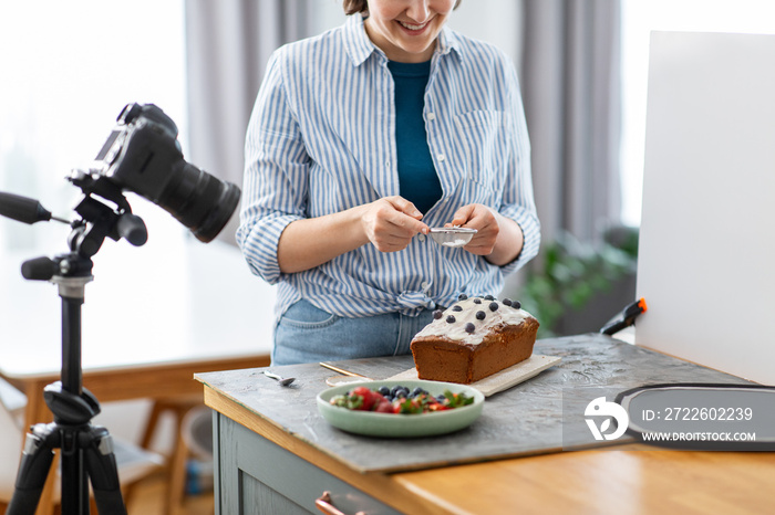 blogging, photographing and people concept - happy smiling female photographer or food blogger with camera pouring powdered sugar to cake in kitchen at home