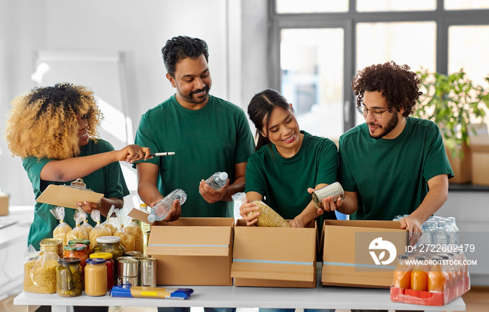 charity, donation and volunteering concept - international group of happy smiling volunteers packing food in boxes according to list on clipboard at distribution or refugee assistance center