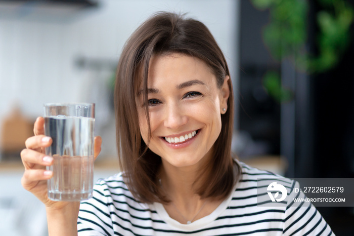 Happy young woman holding glass of water, looks at the camera and smiles friendly. Healthy lifestyle concept