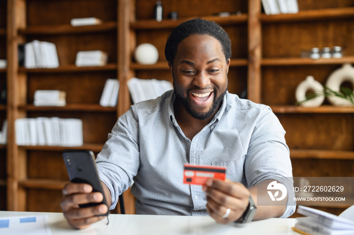 Overjoyed African-American male entrepreneur wearing smart casual shirt holds phone and banking card, making an online order, paying for food delivery in the office, shopping online, e-banking concept