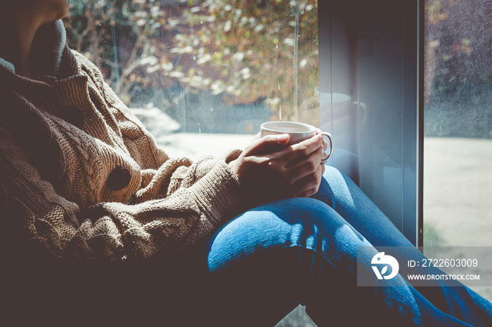 Young brunette woman drinking a cup of tea. Female sitting at home by the window. Cold autumn days. Fall background.