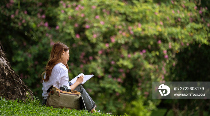 Image of joyful woman holding diary book writing note while sitting on grass in park.