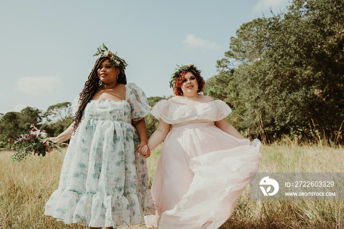 2  plus size friends hold hands and walk in a field wearing dresses