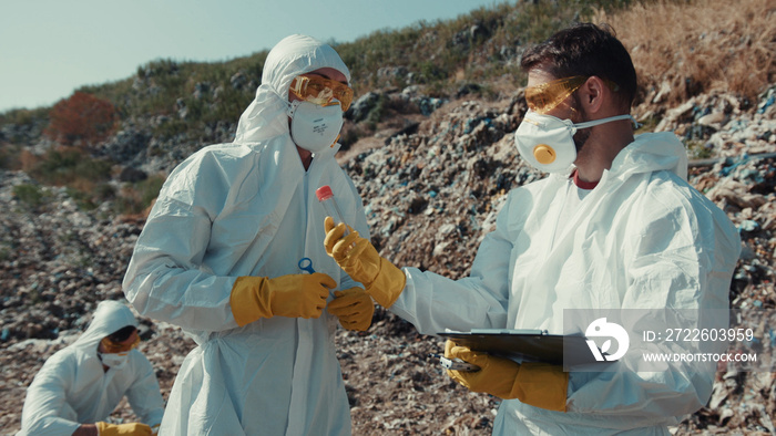 Environmental pollution issue. Team of professional ecologists researching dump in sunny weather. Two scientists examine toxic sample and write down results.