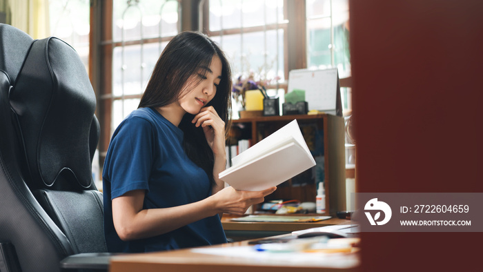 Young adult asian single woman reading a book at home office on day.