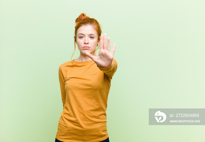 young pretty red head woman looking serious, stern, displeased and angry showing open palm making stop gesture against green wall