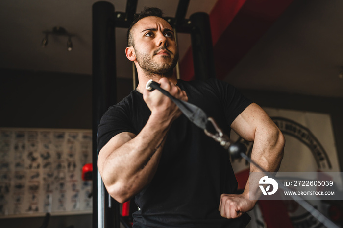 One man young adult caucasian male bodybuilder training arms bicep on the cable machine in the gym holding weight wearing black shirt dark photo real people copy space side view low angle