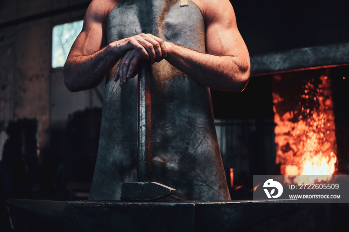 Muscular blacksmith in protective apron at his workshop with hammer in his hands. There are fire at background.