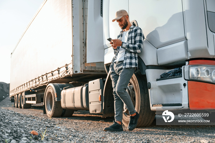 Leaning by the vehicle and using phone. Young truck driver is at daytime