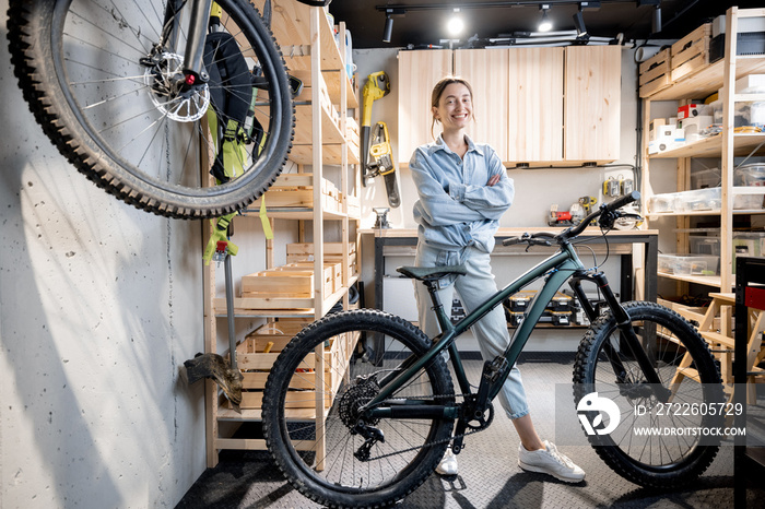 Portrait of a young handywoman standing with a bicycle in the workshop or garage at home. Bicycle repairing and diy concept