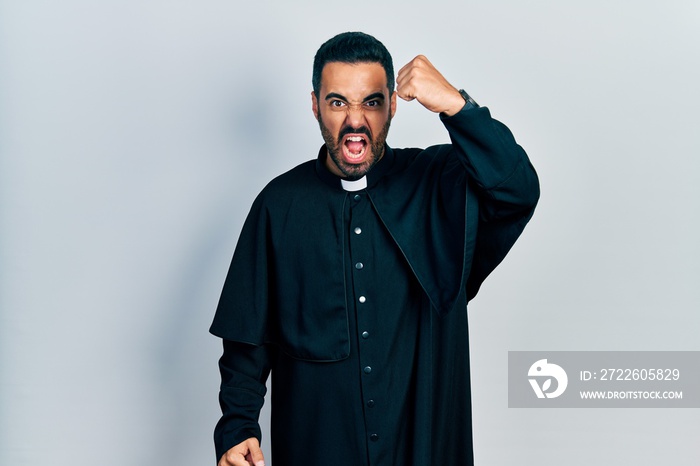 Handsome hispanic man with beard wearing catholic priest robe angry and mad raising fist frustrated and furious while shouting with anger. rage and aggressive concept.