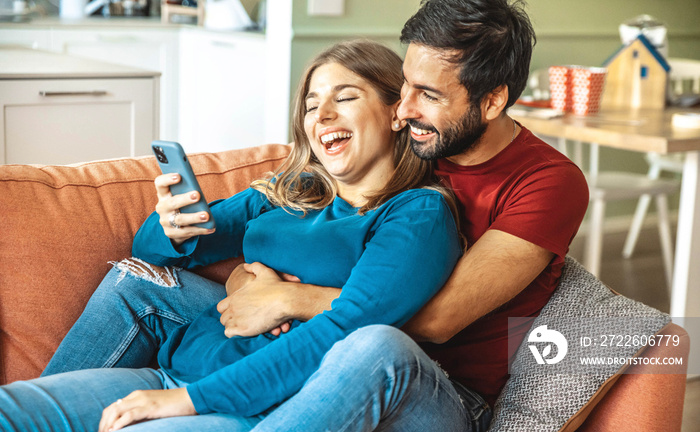 Happy couple watching smart mobile phone device sitting on sofa at home - Husband and wife having fun together using cellphone - Happy lifestyle and technology concept