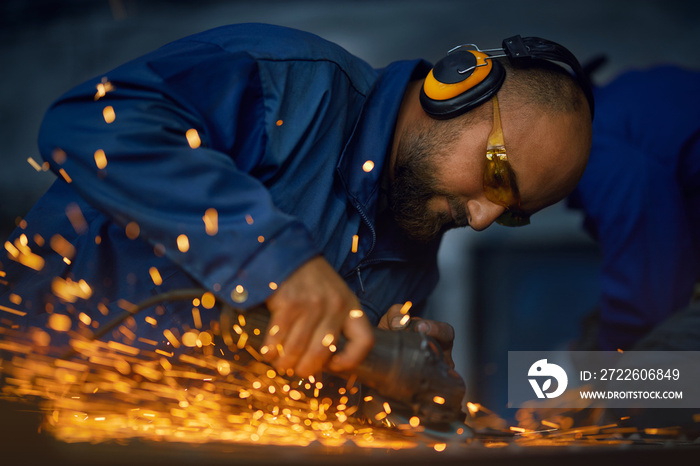 Competent industrial worker in protective clothes, glasses and headphones polishing metal product using angle grinder. Manual work concept.