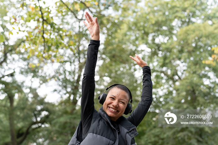 Portrait of smiling woman with headphones raising arms in park