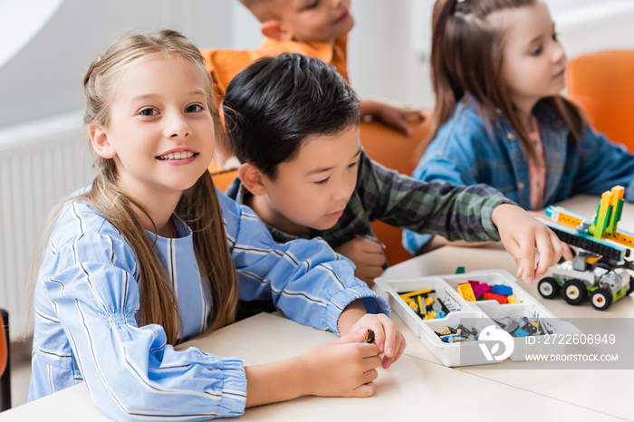 Selective focus of schoolgirl looking at camera near multiethnic friends modeling robot in classroom