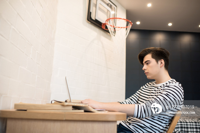 Side view portrait of bored young man browsing internet via laptop sitting at desk in boys room, copy space