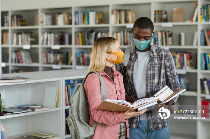 Waist up portrait of two students wearing masks while standing in school library and holding books, copy space