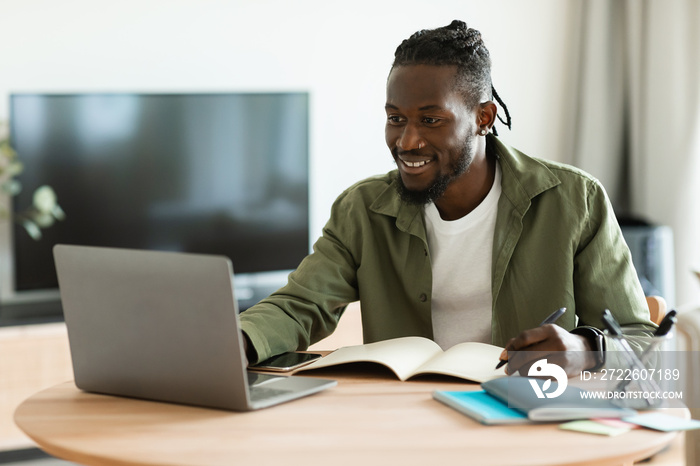 Excited black guy looking for job online, sitting at table, using laptop computer and taking notes, copy space