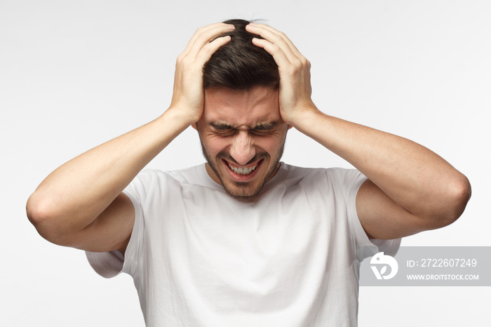 Upset unhappy young man in white t-shirt squeezing head with hands, suffering from headache. People, stress, tension and migraine concept