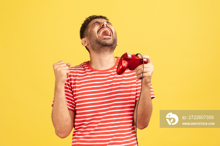 Extremely happy bearded man in striped t-shirt holding hand red joystick pad showing yes gesture sincerely rejoicing, victory in online virtual game. Indoor studio shot isolated on yellow background