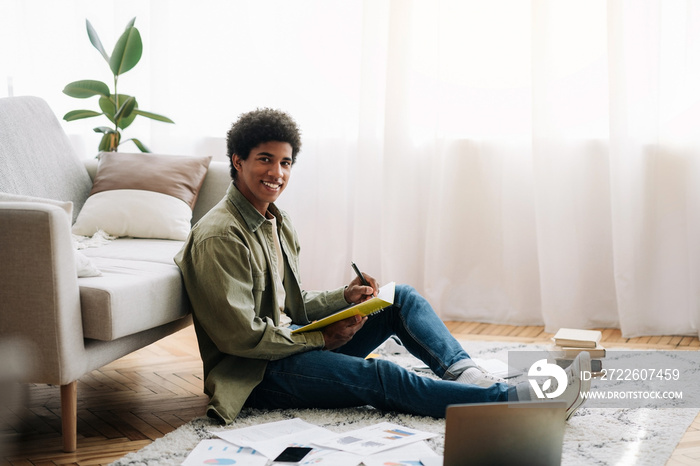 Black teen student with laptop taking notes for difficult exam during online studies at home, free space