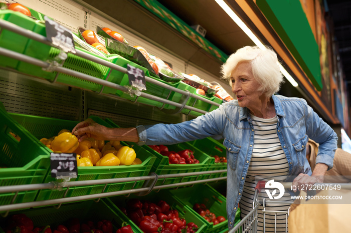 Portrait of modern senior woman with shopping cart  choosing fruits and vegetables in supermarket while enjoying grocery shopping, copy space