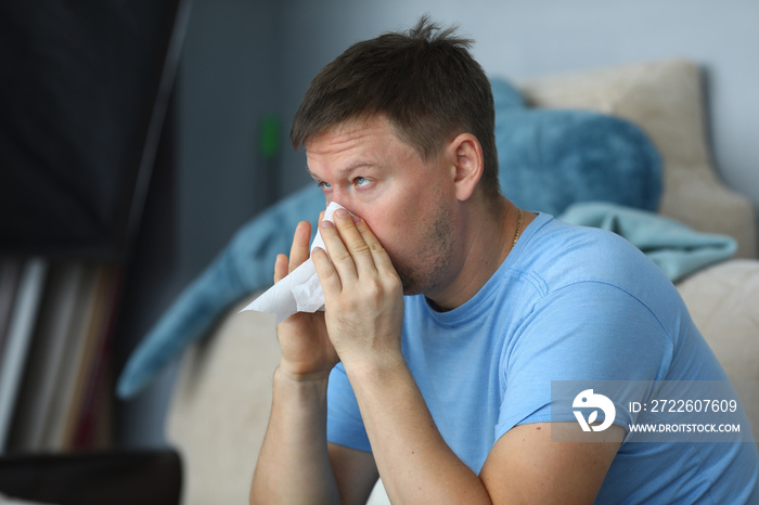 Man blowing his nose in a napkin while sitting at home on sofa portrait