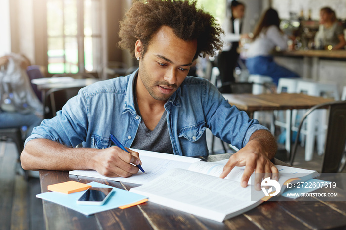 Handsome stylish Afro American university student studying at cafe after lectures, making notes from textbook in copybook, sitting at wooden table with mobile phone with copy space blank screen