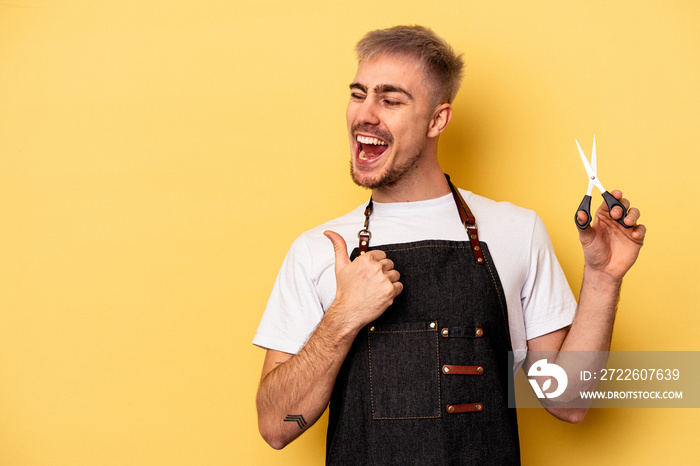 Young caucasian hairdresser man holding scissors isolated on yellow background points with thumb finger away, laughing and carefree.