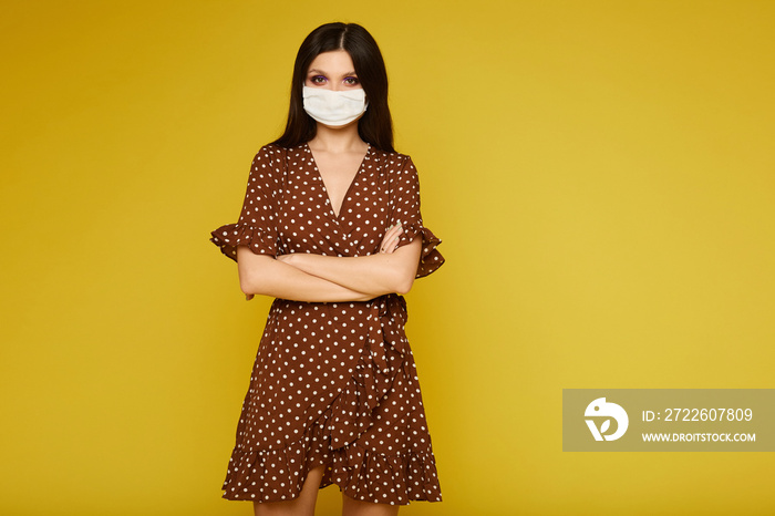 Young woman in dress and medical mask looking at the camera and posing on yellow background, isolated. Model girl protective herself against the flu epidemic, dust allergy, and the virus.