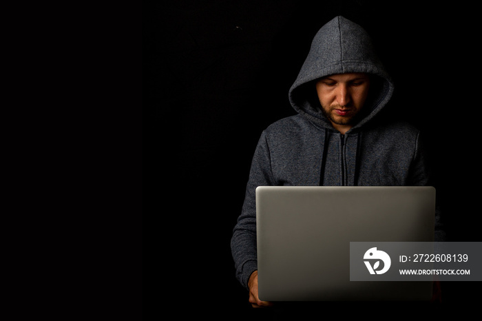 Man in a hood holds a laptop in his hands on a dark background. Concept of cyber rogue, hacker. Banner