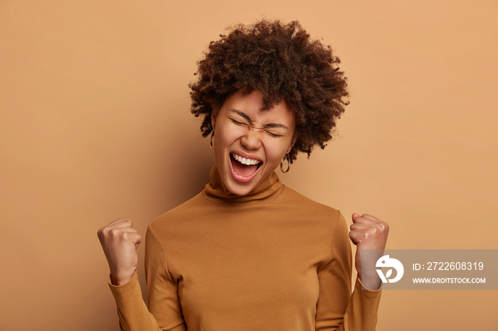 Ambitious lucky triumphing woman celebrates amazing victory with both clenched fists, hears excellent news, exclaims positively hooray, wears brown turtleneck, rejoices over awesome achievement.