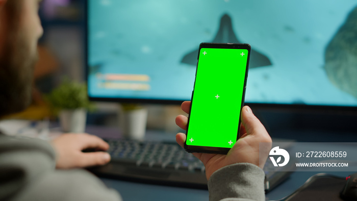 Competitive gamer looking at smartphone with green screen chroma key desktop playing online games on powerful computer during live tournament. Cyber player using phone with mock up isolated display