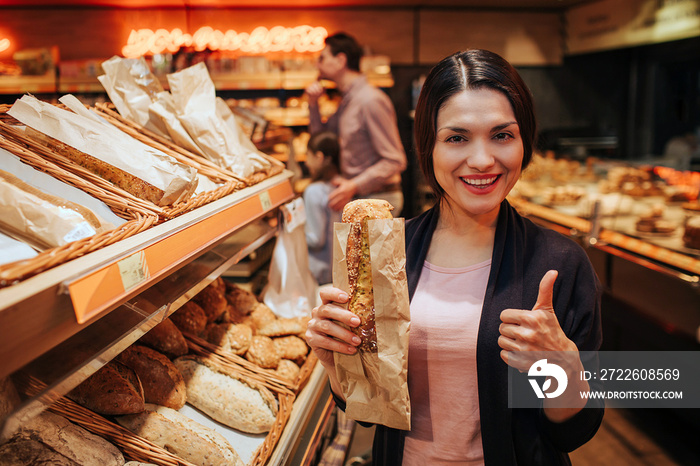 Young parents and daughter in grocery store. Positive cheerful woman hold bread in hands and show big thumb up. Fathr and daughter stand behind and pick up rolls.