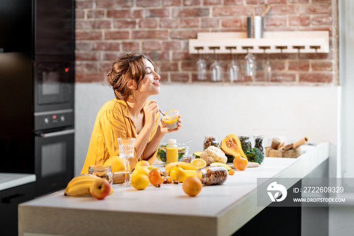 Cheerful woman eating chia pudding at the table full of healthy raw vegetables and fruits on the kitchen at home. Concept of vegetarianism, healthy eating and wellness