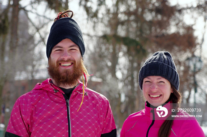 Cheerful couple wearing black hats and pink sports jackets laughing and looking at camera, breathing out steam, winter park behind them. Enjoying outdoor activity together. Concept of fun