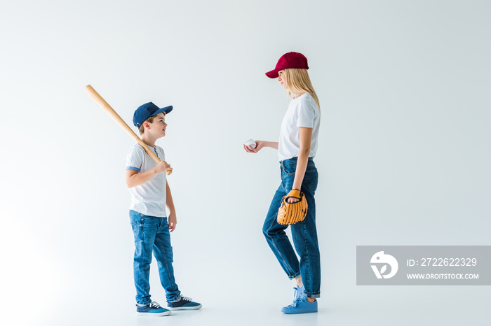 mother and son standing with baseball bat, glove, ball and looking at each other on white